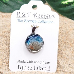 Op de afbeelding: Een zilveren ketting met een ronde glazen hanger die zand en blauwe hars bevat, die de oceaan voorstelt. Het label luidt "K & T Designs The Georgia Collection Made with sand from Tybee Island".