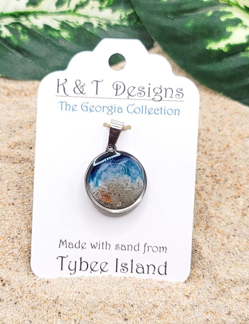 Op de afbeelding: Een zilveren ketting met een ronde glazen hanger die zand en blauwe hars bevat, die de oceaan voorstelt. Het label luidt "K & T Designs The Georgia Collection Made with sand from Tybee Island".