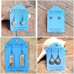 May include: Four pairs of earrings made with sand from Fort Myers, Florida. Each pair is displayed on a blue card with the text "K&T Designs" and "Just Beachy Collection". The earrings are made with different shapes and sizes of sand.