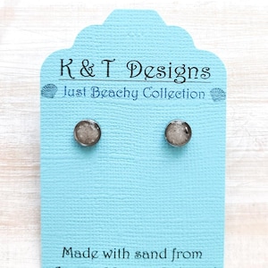 May include: A pair of silver stud earrings with a clear resin top filled with sand from Anna Maria Island. The earrings are displayed on a blue card with the text "K&T Designs Just Beachy Collection" and "Made with sand from Anna Maria Island."