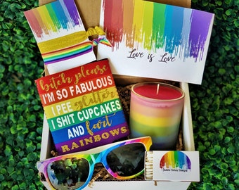 May include: A gift box filled with rainbow-themed items, including a rainbow can cooler with the text "bitch please I'M SO FABULOUS I PEE glitter I SHIT CUPCAKES AND fart RAINBOWS", rainbow sunglasses, a rainbow candle, and a pack of matches with a rainbow design. The box also includes a card with the text "Love is Love" and a rainbow hair tie.