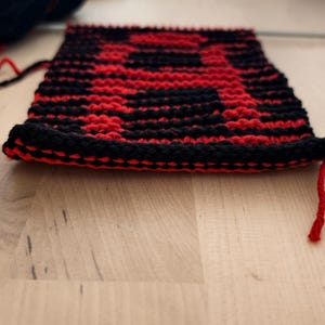 May include: A rectangular piece of double-knit fabric featuring a bold, repeating geometric pattern in vibrant red and deep black yarn. The textile displays a thick, textured construction with visible horizontal rows of stitches and loose yarn ends.