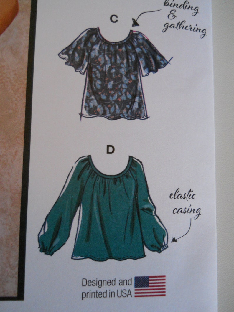 May include: Two illustrations of blouses with different neckline and sleeve styles. The top blouse has a floral print and a binding and gathering detail at the neckline. The bottom blouse is solid green with an elastic casing detail at the sleeve cuffs. The text "Designed and printed in USA" is below the illustrations.