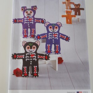 May include: Simplicity sewing pattern number S9417 for making a plush toy bear. The pattern includes instructions for making three different bears, one purple, one blue, and one black. The bears have a skeleton design and are decorated with red and white details. The pattern is for one size.