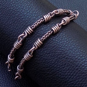 Könnte beinhalten: Zwei Kupferarmbänder mit geflochtenem und Gliederketten-Design. Die Armbänder liegen auf einer schwarzen Lederfläche.