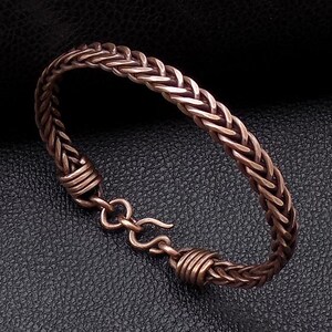 May include: A braided copper bracelet with a hook clasp. The bracelet is made of multiple strands of copper wire that are woven together to create a textured, braided design.