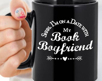 May include: Black ceramic mug with the text "Shhh. I'm on a date with My Book Boyfriend" in white script. The mug has a black handle and a white arrow and heart design.