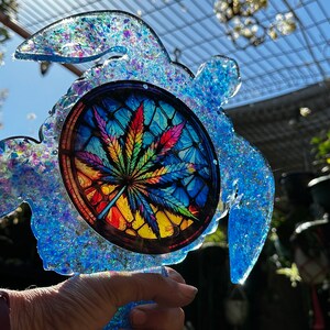 May include: A blue glass turtle with a stained glass design of a marijuana leaf in the centre. The turtle is covered in iridescent glitter.