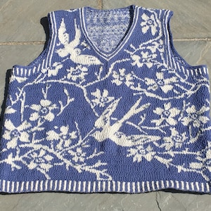 Könnte beinhalten: Eine blaue Strickweste mit einem weißen Blumen- und Vogelmuster. Die Weste hat einen V-Ausschnitt und ist ärmellos.