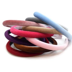 May include: A collection of velvet headbands in a range of colours, such as red, pink, purple, brown, and blue. The headbands are piled together on a white background, highlighting their soft texture and bright colours. These accessories are ideal for adding a touch of style.
