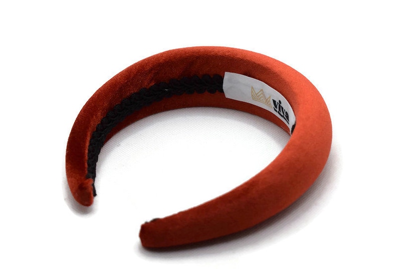 Puede incluir: Una diadema ancha de terciopelo color &oacute;xido con una banda el&aacute;stica negra y una etiqueta blanca que dice "Viva" con un logotipo de corona.