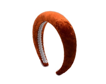 May include: A wide, orange velvet headband with a white trim. The headband is curved and has a smooth, soft texture.