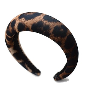 Puede incluir: Una diadema ancha de color marrón y negro con estampado de leopardo. La diadema está hecha de un material suave y acolchado.