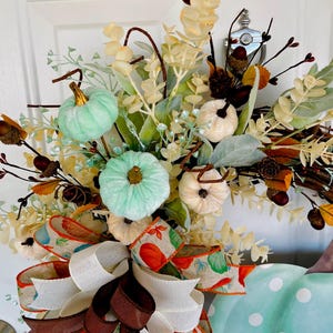 May include: A teal and white polka dot pumpkin wreath with a brown and white ribbon bow. The wreath is decorated with faux greenery, white pumpkins, and teal pumpkins.