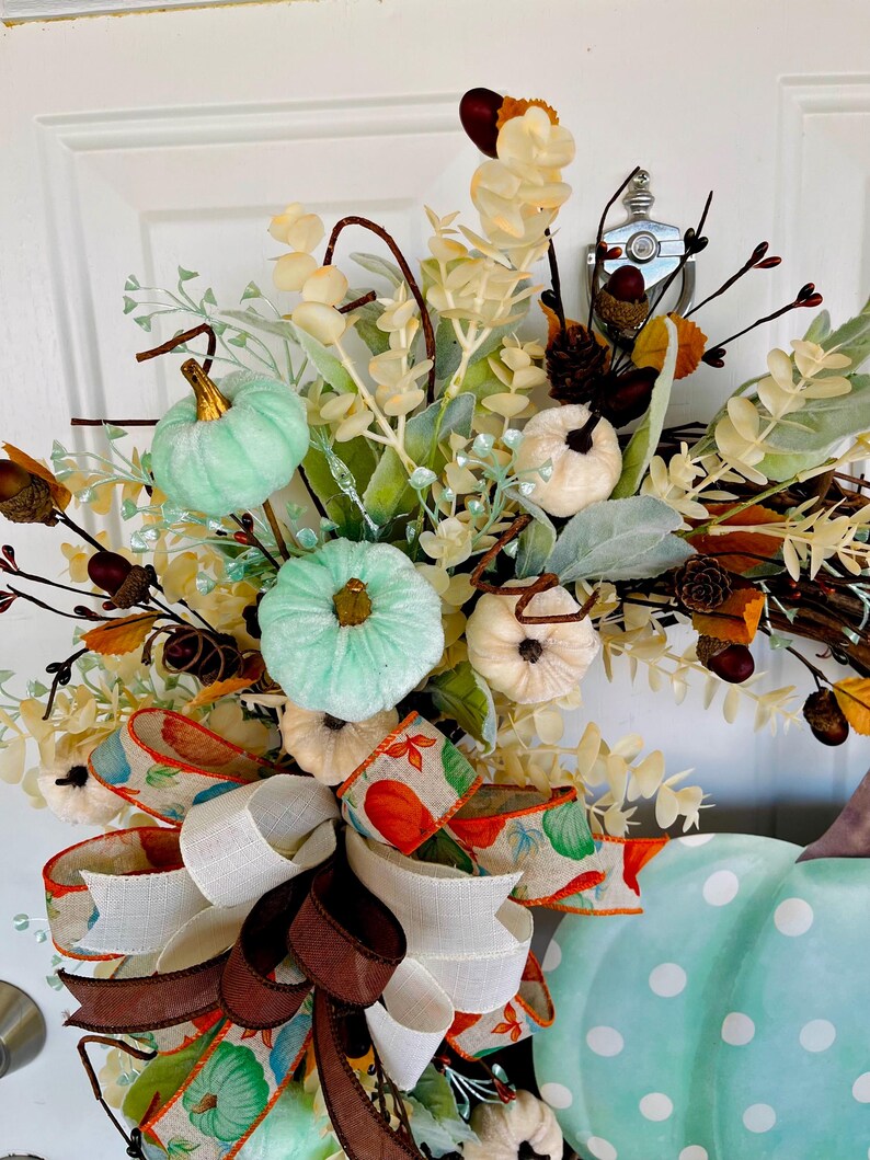 May include: A teal and white polka dot pumpkin wreath with a brown and white ribbon bow. The wreath is decorated with faux greenery, white pumpkins, and teal pumpkins.