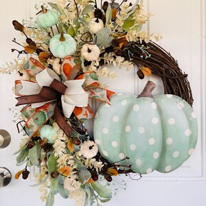 May include: A green and white polka dot pumpkin wreath with brown ribbon and white and green pumpkins. The wreath is made of twigs and has a variety of fall foliage.
