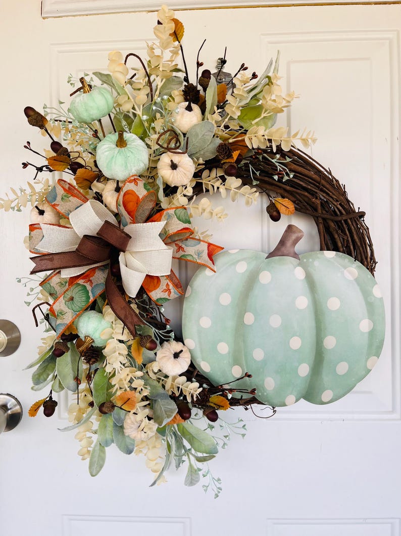 May include: A green and white polka dot pumpkin wreath with brown ribbon and white and green pumpkins. The wreath is made of twigs and has a variety of fall foliage.