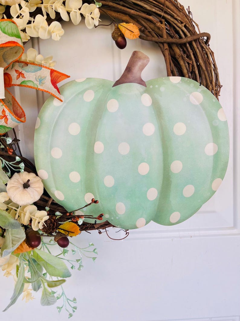 May include: A green and white polka dot pumpkin wreath with a brown stem. The wreath is made of grapevine and has a variety of fall foliage, including leaves, berries, and pine cones.