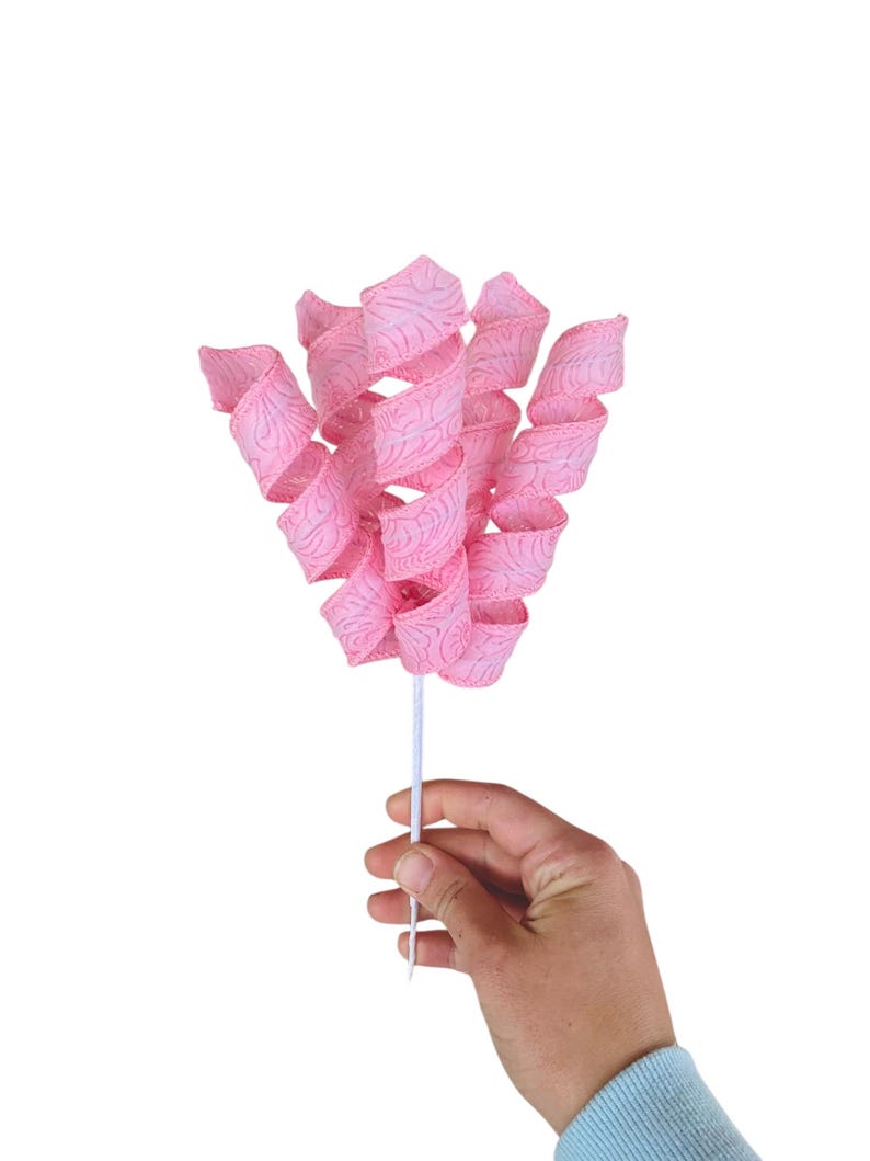 May include: A pink ribbon with a swirl design is attached to a white stick. The ribbon is curled and twisted to create a flower-like shape.