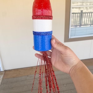 May include: A red, white, and blue decorative rocket with glitter accents. The top is red, followed by a white section, and a blue base. Red glitter strands extend from the bottom, creating a festive look.