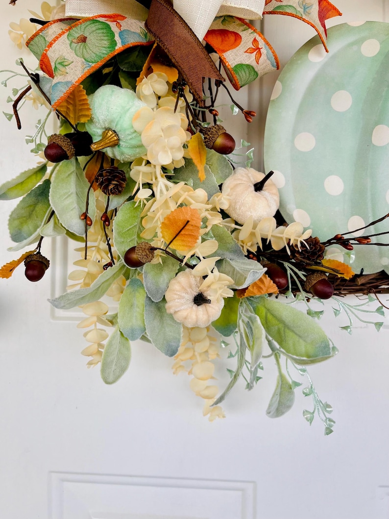 May include: A green and white polka dot pumpkin wreath with a brown ribbon, green and white pumpkins, and fall foliage.