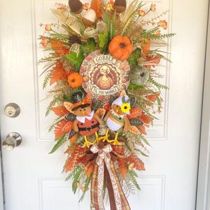 May include: Thanksgiving-themed door wreath with a turkey and pumpkin design. The wreath features a circular sign that says "Gobble Til You Wobble". The wreath is decorated with orange, brown, and green leaves, acorns, and pumpkins.