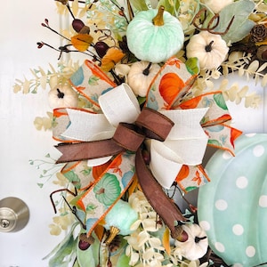 May include: A fall-themed wreath with a large, light blue pumpkin with white polka dots. The wreath is decorated with a variety of fall foliage, including eucalyptus, berries, and acorns. A large bow made of brown, white, and orange ribbon with a pumpkin print is tied at the top of the wreath.