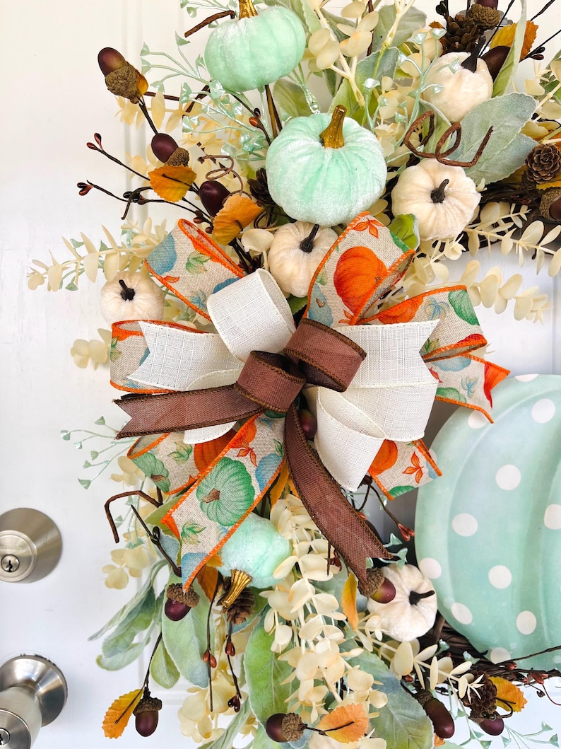 May include: A fall-themed wreath with a large, light blue pumpkin with white polka dots. The wreath is decorated with a variety of fall foliage, including eucalyptus, berries, and acorns. A large bow made of brown, white, and orange ribbon with a pumpkin print is tied at the top of the wreath.