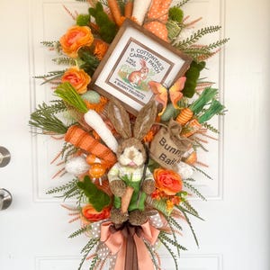 May include: Easter-themed door wreath with a bunny and carrot theme. The wreath features a plush bunny holding a carrot, orange roses, faux carrots, and a sign that reads "P. Cottontail's Carrot Patch". The wreath is decorated with a bow and greenery.
