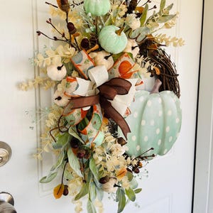 May include: A green and white pumpkin wreath with a brown and white ribbon. The wreath is decorated with fall foliage and berries.