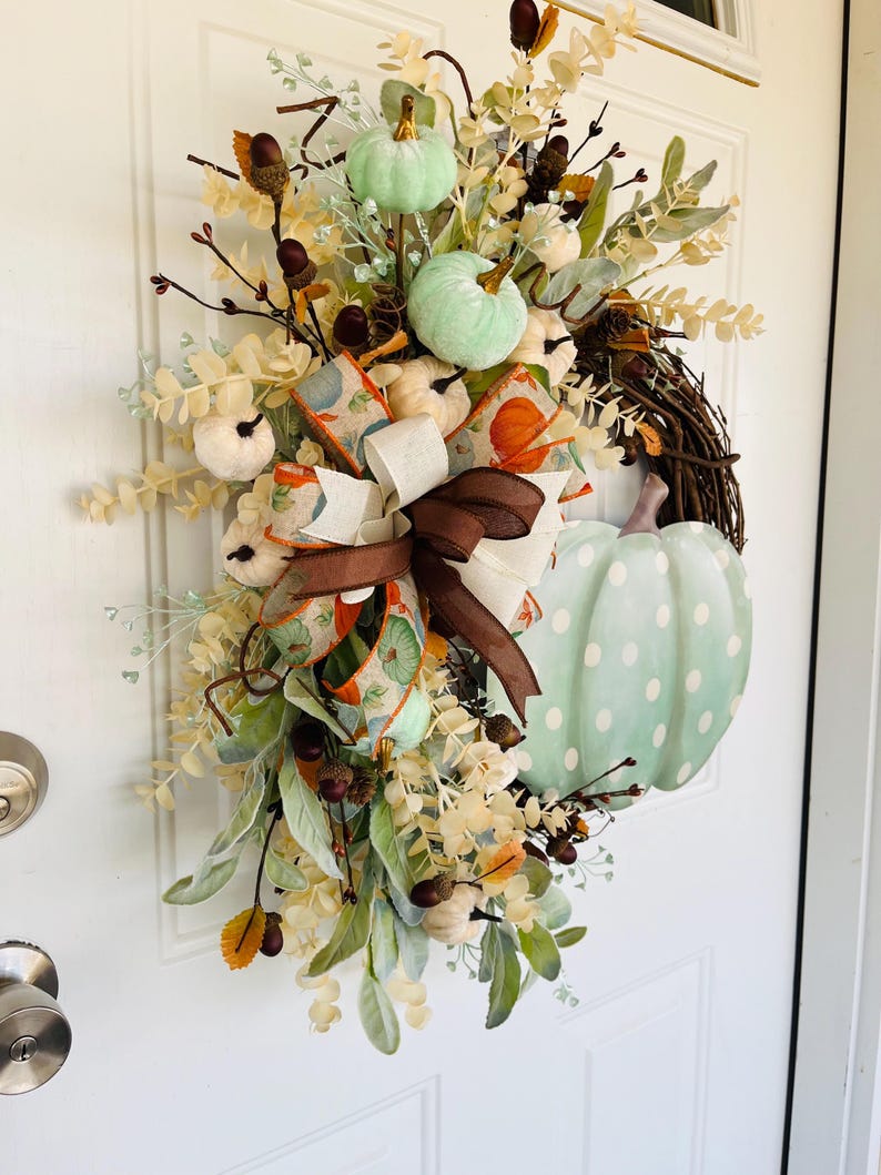May include: A green and white pumpkin wreath with a brown and white ribbon. The wreath is decorated with fall foliage and berries.