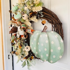 May include: A green and white polka dot pumpkin with a brown stem is featured on a grapevine wreath. The wreath is decorated with fall foliage, including white and green pumpkins, brown berries, and a brown and white ribbon.