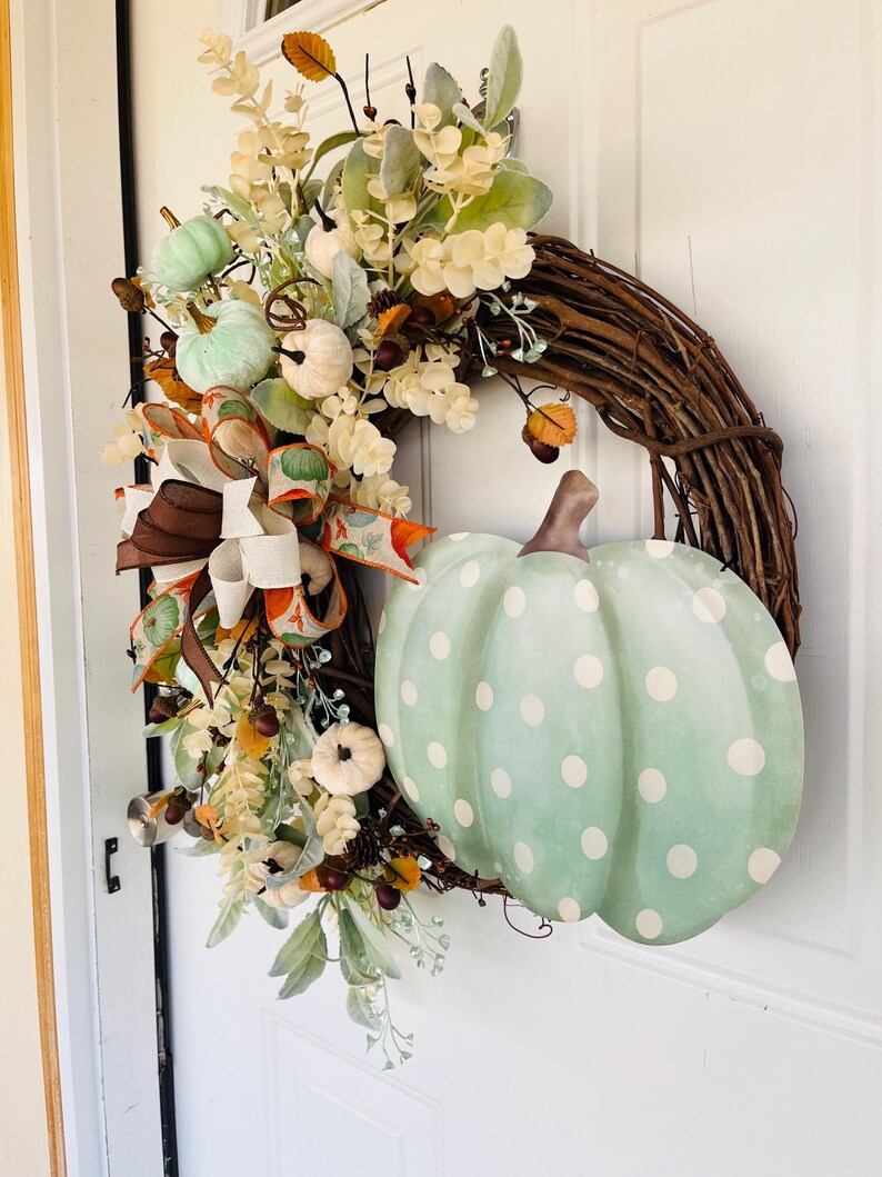 May include: A green and white polka dot pumpkin with a brown stem is featured on a grapevine wreath. The wreath is decorated with fall foliage, including white and green pumpkins, brown berries, and a brown and white ribbon.
