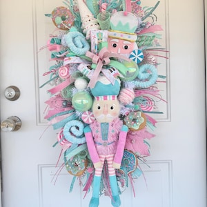May include: A decorative Christmas wreath in pastel shades, featuring a nutcracker doll, donuts, and sweets. The wreath is embellished with pink, blue, and white ribbons, baubles, and other festive items. It is hanging on a white door.