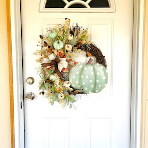 May include: A green and white polka dot pumpkin with a brown stem is featured on a wreath with a brown bow and white and green pumpkins. The wreath is hanging on a white door.