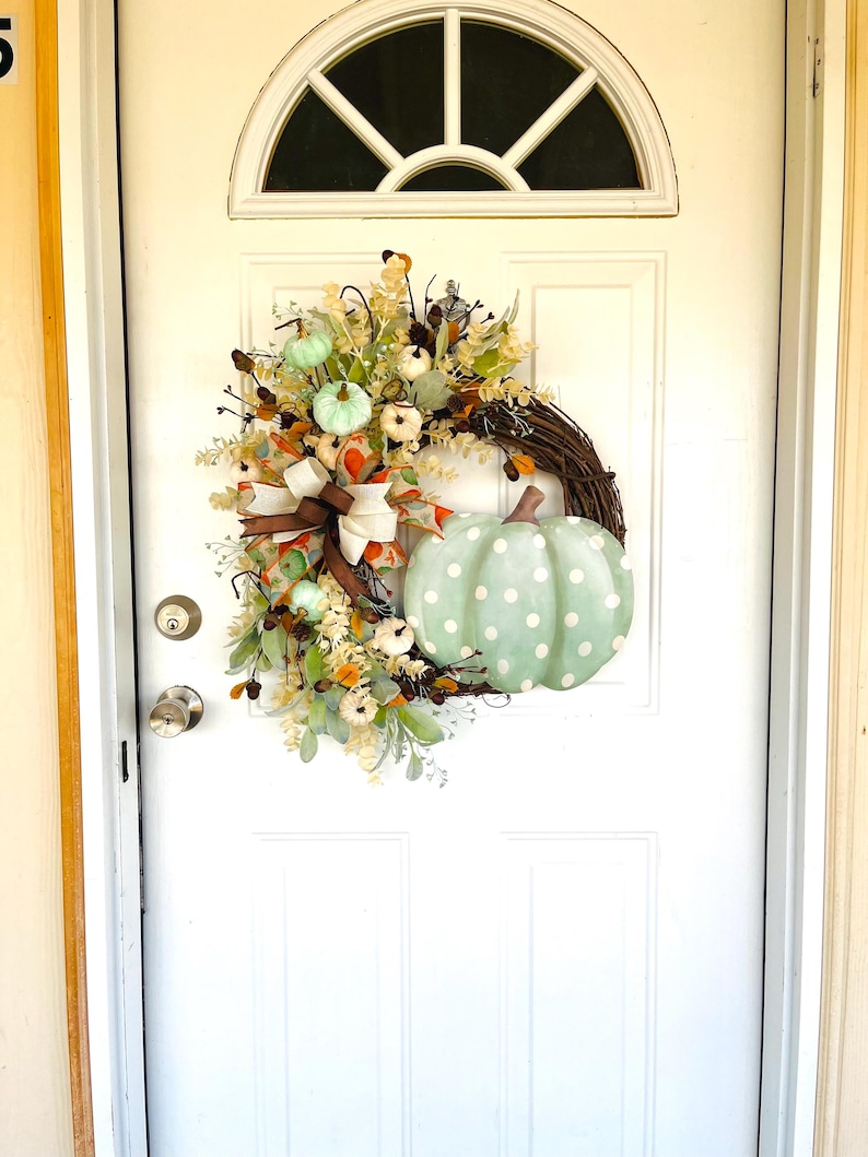 May include: A green and white polka dot pumpkin with a brown stem is featured on a wreath with a brown bow and white and green pumpkins. The wreath is hanging on a white door.
