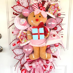 May include: A decorative holiday wreath featuring a plush gingerbread figure holding a light blue gift box. The wreath is adorned with pink and white ornaments, miniature gingerbread houses, candy-themed accents, and a large pink ribbon bow.