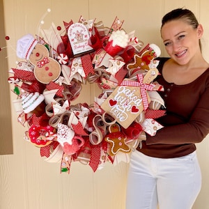 May include: A decorative holiday wreath featuring red and white mesh ribbons, gingerbread man ornaments, a snowman wearing a chef hat, and a wooden sign that reads Baked with Love. The wreath includes miniature mugs, donuts, and candy accents.