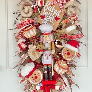 May include: A festive Christmas wreath with a baking theme. It features a wooden nutcracker, gingerbread men, and a sign that says "Baked with Love." The wreath is decorated with red and white accents, candy canes, and a Santa hat.
