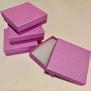 May include: Several stacked, square, light pink gift boxes. One box is open, revealing white padding inside. The boxes are arranged on a light-colored surface, with the open box slightly angled.