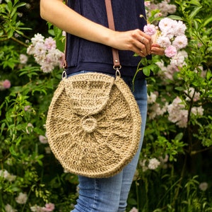 May include: A round, beige crochet bag with a flap closure and a brown leather strap. The bag has a textured, woven design and is worn over the shoulder. The background features green foliage and pink flowers.