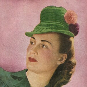 May include: A woman with brown hair is wearing a green knitted hat with pink and purple pom-poms. She is wearing a green dress and looking to the side.