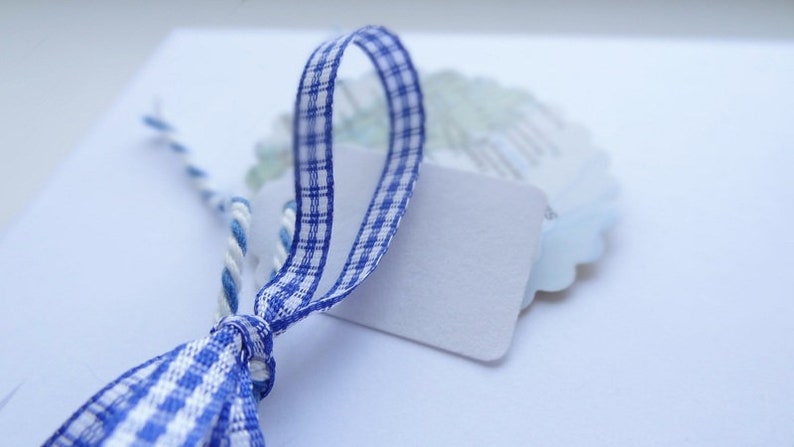 May include: A blue and white checked ribbon tied around a blank white gift tag. The tag is attached to a white and blue twine.