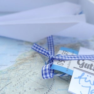 May include: A blue and white checked ribbon tied around a blue and white gift tag with the text "Gutschein f&uuml;r Jer" on a white card. The gift tag is on top of a map.