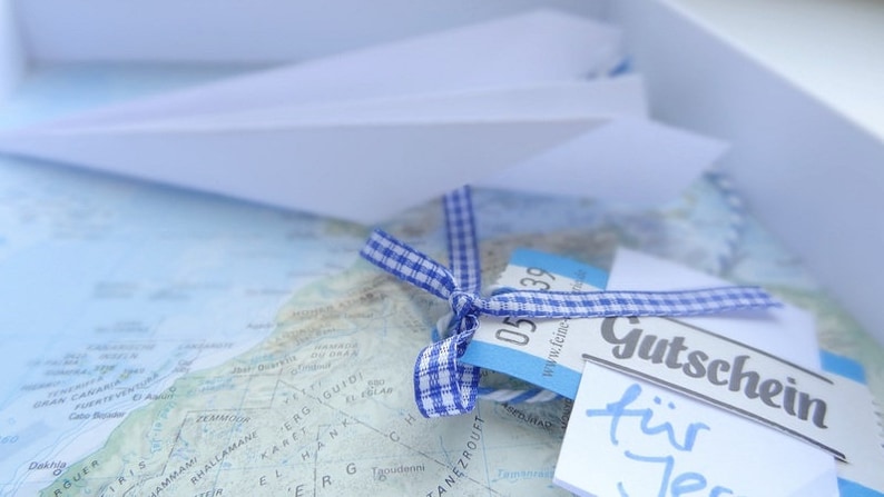 May include: A blue and white checked ribbon tied around a blue and white gift tag with the text "Gutschein f&uuml;r Jer" on a white card. The gift tag is on top of a map.