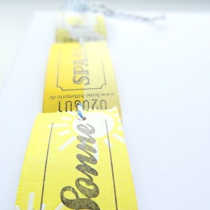May include: Yellow paper tickets with the text "Sommer" and "Spain" printed on them. The tickets are tied together with a blue string.