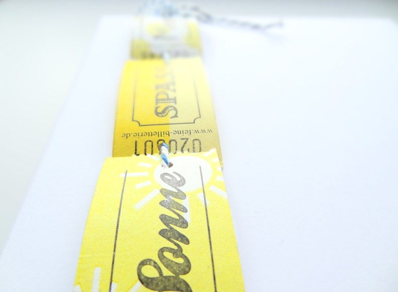 May include: Yellow paper tickets with the text "Sommer" and "Spain" printed on them. The tickets are tied together with a blue string.