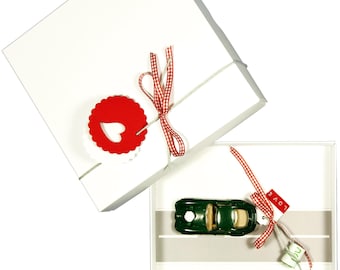 Può includere: Una scatola regalo bianca con un nastro a quadretti rossi e bianchi e un'etichetta rossa a forma di cuore. All'interno, un'auto cabriolet giocattolo verde scuro su una grafica stradale, con un'etichetta rossa con la scritta LOVE e rotoli di carta.