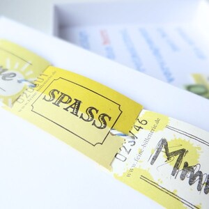 May include: Yellow paper tickets with the words "Sommer" and "SPASS" printed on them. The tickets have a blue string attached to them and are on a white surface.