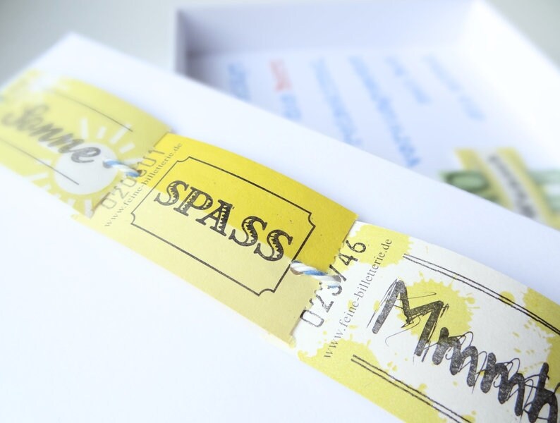 May include: Yellow paper tickets with the words "Sommer" and "SPASS" printed on them. The tickets have a blue string attached to them and are on a white surface.
